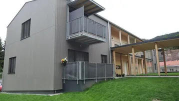 Modernes Gebäude mit grauer Fassadenverkleidung, Balkonen und einer Grünfläche davor.