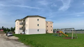 Neubau-Mehrfamilienhaus mit Balkonen und einem Spielplatz auf einer grünen Wiese unter blauem Himmel.