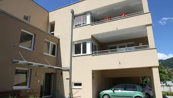 Modernes Mehrfamilienhaus mit beiger Fassade, Balkonen und überdachtem Stellplatz für Fahrzeuge.