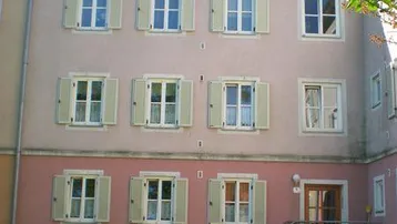 Mehrstöckiges Wohngebäude mit hellrosa Fassade, grünen Fensterläden und einem kleinen Vorgarten.