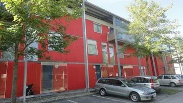 Modernes Wohngebäude mit roter Fassade, Balkonen und Außenparkplätzen im Grünen.