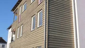 Moderne Gebäudefassade mit beiger Holzverkleidung, großen Fenstern und einem französischen Balkon unter blauem Himmel.