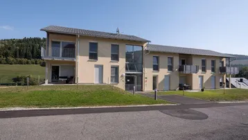 Modernes Mehrfamilienhaus mit Balkonen und großen Fenstern, eingebettet in eine grüne Landschaft.