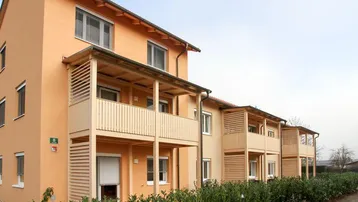 Modernes Mehrfamilienhaus mit orangefarbener Fassade, Holzverkleidung an den Balkonen und grüner Hecke.