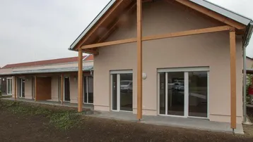 Neubau mit heller Fassade, großen Fenstern und sichtbarer Holzbalkenstruktur im Außenbereich.