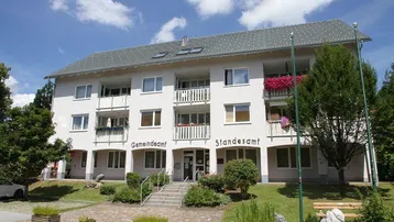 Mehrfamilienhaus mit weißer Fassade, Satteldach und Balkonen in grüner Umgebung.