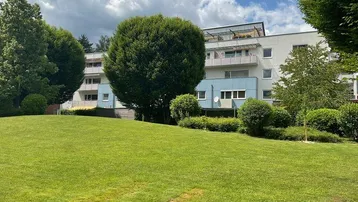 Mehrfamilienhaus mit großer Grünfläche und Bäumen im Vordergrund unter bewölktem Himmel.