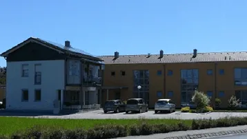 Mehrfamilienhaus mit zwei Gebäudeteilen, Photovoltaikanlage auf dem Dach und Parkplätzen davor.