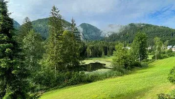 Panoramablick auf eine grüne Landschaft mit Bergen, einem kleinen See und üppigen Bäumen unter bewölktem Himmel.