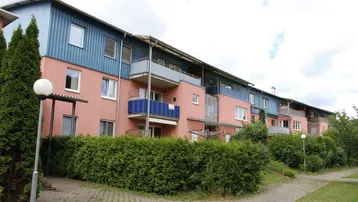Mehrfamilienhaus mit rosa Fassade, blauer Verkleidung und mehreren Balkonen, umgeben von gepflegten Grünflächen.