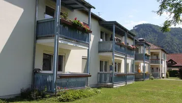 Mehrfamilienhaus mit mehreren Balkonen und Blick auf die umliegende Berglandschaft, umgeben von einer grünen Rasenfläche.
