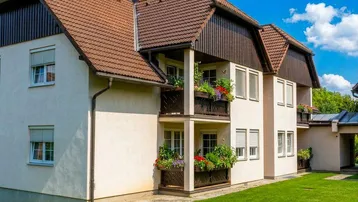 Mehrfamilienhaus mit heller Fassade, braunem Dach und blühenden Balkonen.