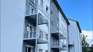 Moderne Fassade eines Mehrfamilienhauses mit mehreren Balkonen, großen Fenstern und Rollläden unter blauem Himmel.