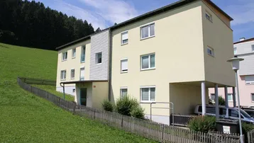 Mehrfamilienhaus mit gelber Fassade, umgeben von grünen Hügeln und Bäumen unter blauem Himmel.