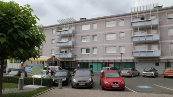 Mehrstöckiges Gebäude mit Balkonen und Rollläden, davor ein Parkplatz mit mehreren Autos und Bäumen.