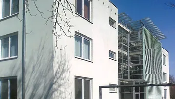 Moderne Gebäudefassade mit heller Putzfassade, vielen Fenstern und einem markanten Glas-Stahl-Anbau.