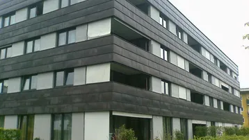 Modernes Mehrfamilienhaus mit dunkler Fassadenverkleidung, großen Fenstern und Rollläden, umgeben von Grünflächen.