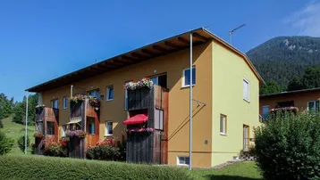 Modernes Mehrfamilienhaus mit gelber Fassade, Balkonen und Bergblick in grüner Umgebung.