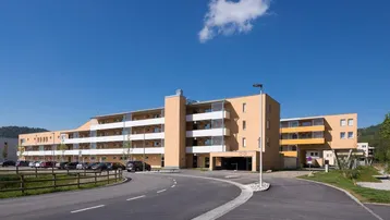 Modernes Mehrfamilienhaus mit mehreren Balkonen und Außenparkplätzen unter blauem Himmel an einem sonnigen Tag.