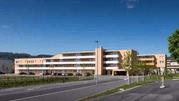 Modernes Mehrfamilienhaus mit hellbeiger Fassade, zahlreichen Balkonen und Außenparkplätzen unter blauem Himmel.