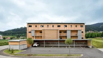 Modernes Mehrfamilienhaus mit Holzfassade, Balkonen und Carport, umgeben von grüner Landschaft.