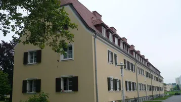 Gelbes Mehrfamilienhaus mit Mansarddach und roten Ziegeln, umgeben von Bäumen und Grünflächen.