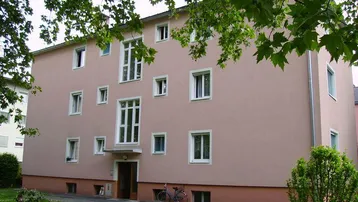 Mehrfamilienhaus mit rosafarbener Fassade und vielen Fenstern, umgeben von Grünflächen.