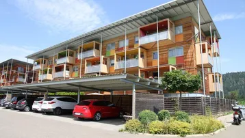 Modernes Mehrfamilienhaus mit Holzfassade, farbigen Balkonen und überdachten Stellplätzen im Grünen.