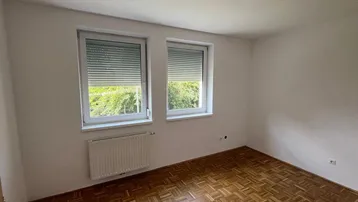 Helles Zimmer mit Parkettboden, weißen Wänden und zwei Fenstern mit Blick ins Grüne.