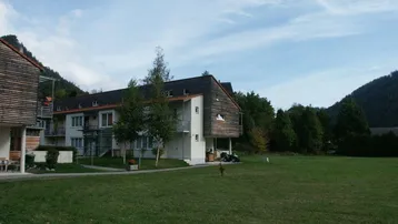 Modernes Wohngebäude mit Holzfassade, umgeben von einer weitläufigen Grünfläche und Bergen im Hintergrund.