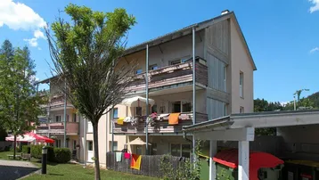 Mehrfamilienhaus mit Balkonen, umgeben von Grünflächen und Bäumen unter blauem Himmel.