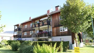 Mehrfamilienhaus mit Holzfassade, Balkonen und gepflegtem Gartenbereich, umgeben von grüner Natur und Bergen.