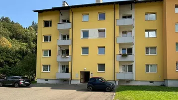 Gelbes Mehrfamilienhaus mit mehreren Balkonen, Parkplätzen und angrenzendem Waldgebiet.