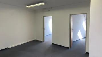Leerer Büroraum mit dunklem Teppichboden, weißen Wänden und zwei offenen Türen zu weiteren Räumen.