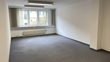 Leerer Büroraum mit Teppichboden, großen Fenstern, Deckenleuchten und Jalousien.
