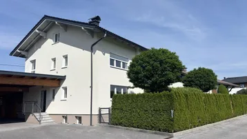 Modernes Einfamilienhaus mit heller Fassade, Carport und gepflegter Hecke im Vorgartenbereich.