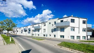 Moderne Wohnanlage mit weißer Fassade und grauen Balkonen entlang einer Straße unter blauem Himmel.