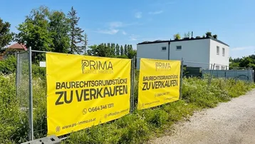 Zwei gelbe Werbebanner für Baugrundstücke an einem Zaun vor einem modernen Gebäude.