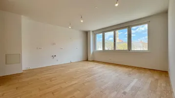 Helles, leeres Wohnzimmer mit Parkettboden und großen Fenstern, die viel Tageslicht hereinlassen.
