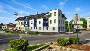 Modernes Mehrfamilienhaus an einer Kreuzung mit blauen Balkonen und gepflegter Umgebung.