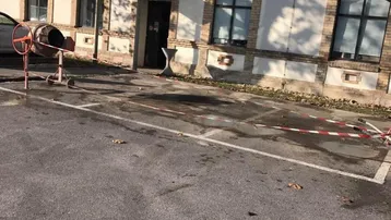 Historisches Gebäude mit Ziegelfassade und Bogenfenstern, umgeben von einem Parkplatz mit Baustellenelementen.