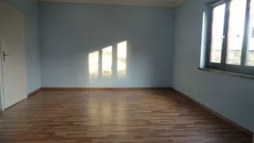 Helles Zimmer mit blauen Wänden und Laminatboden, durch ein Fenster fällt Sonnenlicht herein.