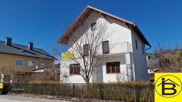 Weißes Einfamilienhaus mit Satteldach und kleinem Balkon, umgeben von Natur und Bäumen.