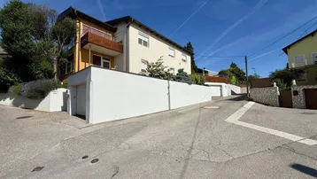 Wohnhaus an einer Hanglage mit Balkon und weißer Stützmauer unter blauem Himmel.
