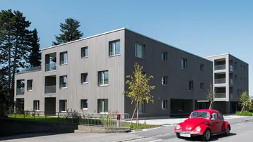 Modernes Mehrfamilienhaus mit Holzfassade und Balkonen an einer ruhigen Straße mit einem roten Käfer.