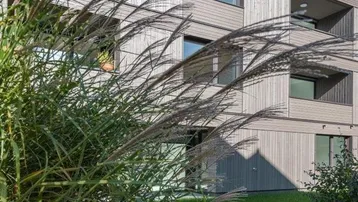 Modernes Gebäude mit Holzfassade und großen Fenstern, umgeben von grüner Vegetation und Rasenfläche.