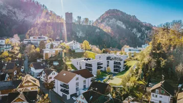 Luftaufnahme einer modernen Wohnanlage in einer bergigen Landschaft mit umliegenden Häusern und Burgruine.