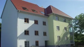 Modernes Mehrfamilienhaus mit weißer und hellgrüner Fassade, rotem Ziegeldach und verglastem Treppenhaus.