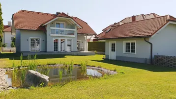 Großzügiger Gartenbereich mit Teich und zwei Wohnhäusern unter blauem Himmel.