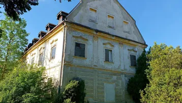 Altes, verputztes Gebäude mit mehreren Fenstern und Mansarddach in ländlicher Umgebung.